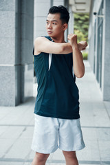 Young Asian guy in sportswear doing arm exercise and looking away while working out near building on street