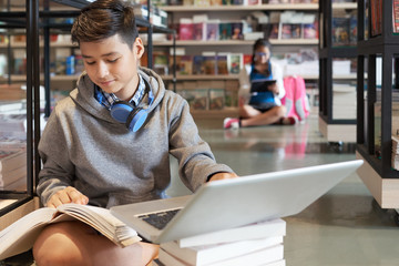 Teenager with headphones reading book and using laptop in library of school