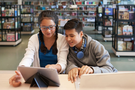 Little girl and boy surfing tablet while sitting at table in school library and smiling