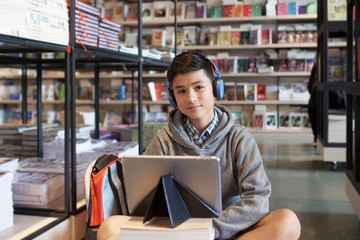 Young student boy sitting on library floor with backpack tablet in headphones looking at camera smiling on bookshelves background