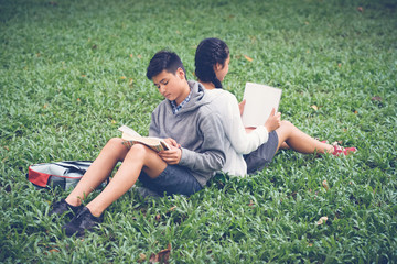 Naklejka na ściany i meble Asian school girl and boy sitting on green grass back to back and reading textbooks
