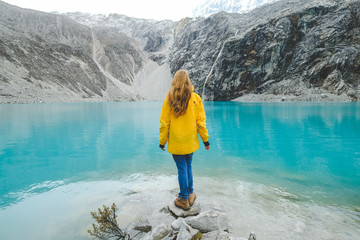 Laguna 69, Peru. A girl stands with her back against a blue lake in a yellow jacket in jeans