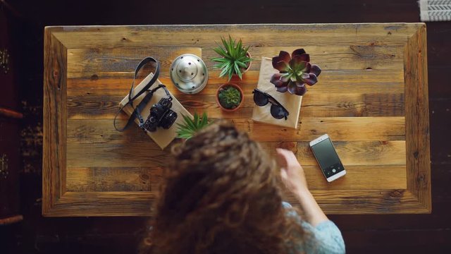 Top view of young woman placing camera, plants and sunglasses on table then taking picture with smartphone creating flay lay photographs. Design and modern technology concept.