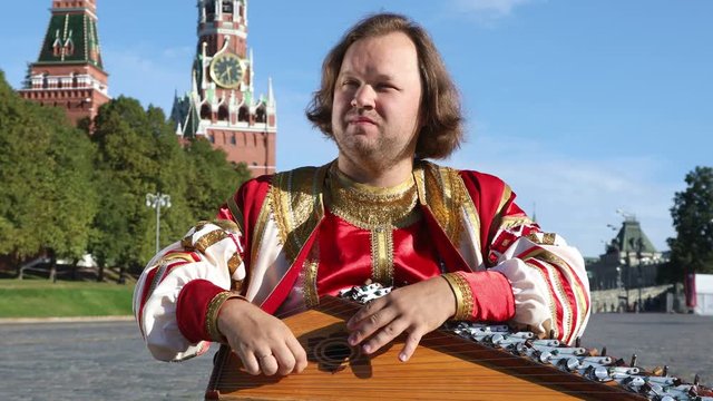 The musician in traditional Russian clothes plays an old Russian musical instrument gusli on the background of the Kremlin on red Square. Moscow, Russia
