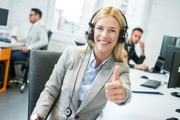 Friendly blonde female telephone worker with headset showing thumb up in office