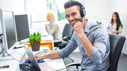 Smiling friendly young call center executive with headset working on computer in office
