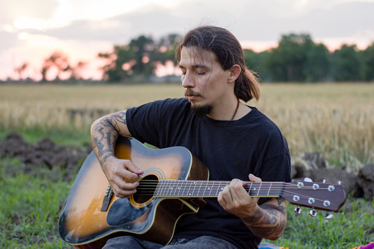 Young tattooed male play on acoustic guitar in summer fields during beautiful sunset	