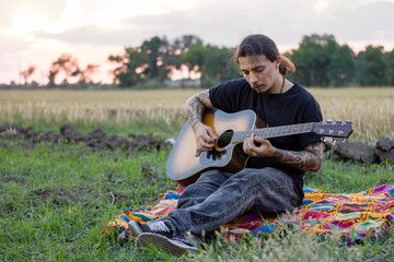 Naklejka na ściany i meble Young tattooed male play on acoustic guitar in summer fields during beautiful sunset 
