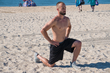 Young healthy athletic man stretching after cardio run workout on sandy beach during hot summer time weather along ocean background. Important stretch muscles after exercise to prevent cramps