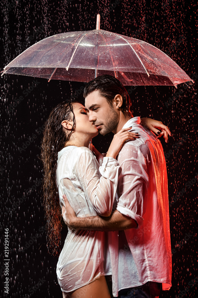 Wall mural side view of romantic couple in white shirts with umbrella standing under rain on black backdrop