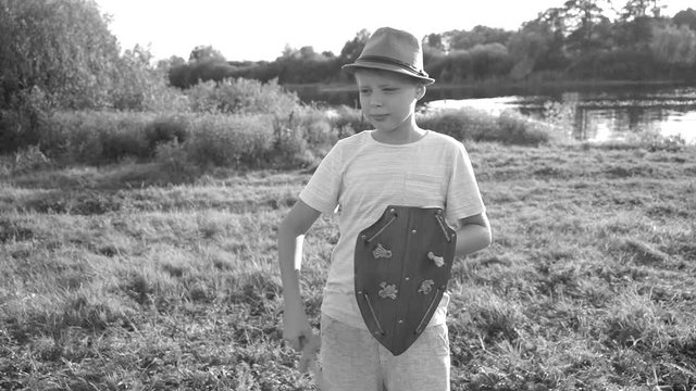 Young white kid playing with wooden toys outside in countryside meadow on sunny sunset summer evening. Boy holds sword and shield. Black and white real time full hd video footage.