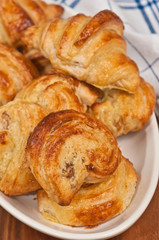Top view, close up of six freshly baked, homemade golden, raisin cream croissant rolls in an oval, white dish with a white and blue striped towel , on a wood cutting board