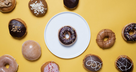 Donuts (doughnuts) on a yellow background.  Classic chocolate ring donut with sprinkles on a white plate surrounded by other donuts on a yellow background