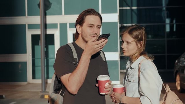 Leisure, technology, communication and people concept - young man and woman using voice command recorder helper or calling on smartphone at street, students voice recognition message with coffee