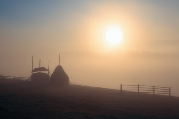 The bright disk of the sun illuminates with a warm orange light the mountain meadow where loose stacked hays built around a central pole stand, which are shrouded in a light mist.
