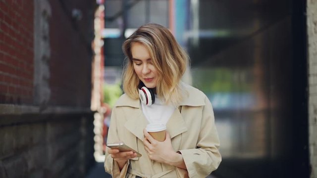 Slow motion of pretty young lady holding smartphone touching screen and drinking to-go coffee outdoors. Urban lifestyle, take away drinks and modern technology concept.