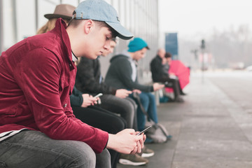 A young adult uses his smartphone at the bus stop. Other people do the same on the blurred background. Concept of young people using mobile phones.