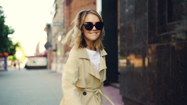 Slow motion portrait of smiling young woman in trendy coat walking in the street, turning to camera and looking at camera. Cheerful people, city and urban lifestyle concept.