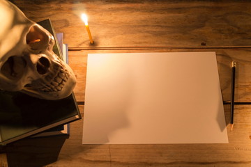 Human skull with candle light on the book and white paper with pencil on wooden table, Decorated for Halloween Theme with copy space.