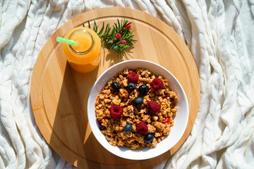 Top horizontal view on healthy breakfast granola fresh berries in white bowl and glass of fresh orange juce on round wooden desk on cozy knitted white plaid in bed.