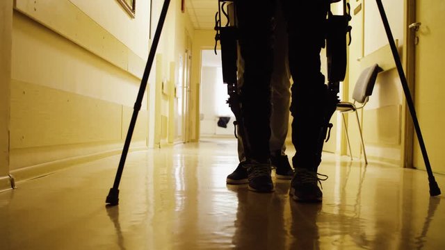 Close up silhouette of the legs of disable man in in the robotic exoskeleton walking through the corridor of the rehabilitation clinic. Rear view