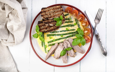 Omelette with asparagus and basil. Homemade ham, cherry tomatoes, toast. Wooden light background. Top view
