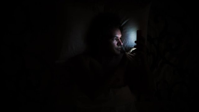 Man in bed turns on phone. Using smartphone in bed due to insomnia health issue. Waking up and using cell phone on pillow in darkness.