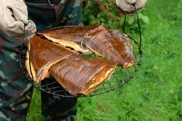 The cooked fish is on a round grill in male hands. The hot-smoked halibut and the flounder are in outdoors. The man is working mittens and camouflage trousers holding seafood.