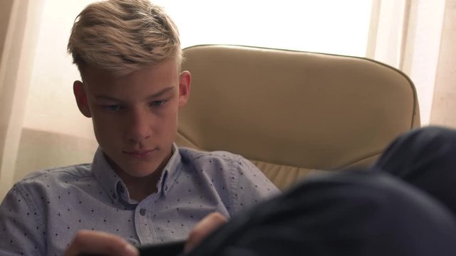 Young boy is sitting with his legs tucked in a chair and clicking on the phone