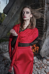 Beautiful fashion model girl wearing a red long skirt and a red shawl stands near the burning brick walls of ruins of collapsed building.
