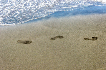 traces of human feet on black sand near the water on the beach. Background texture.