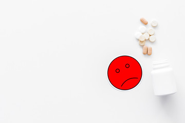 Illness. Drug intake. Pills falling out of jar near sad face emoji on white background top view copy space