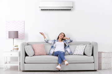 Young woman switching on air conditioner while sitting on sofa near white wall