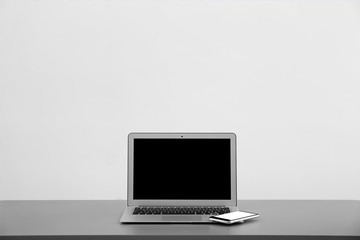 Modern laptop with blank screen and mobile phone on table against light background