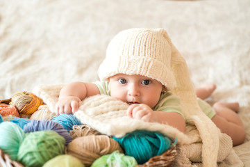 cute baby in a white knitted cap with a bumbon on a wicker basket. Multi-colored knitting threads....