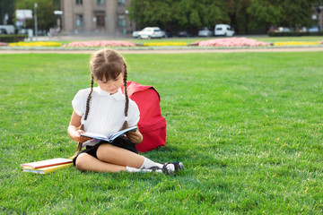 Cute girl with school stationery reading book on green lawn outdoors