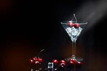 Close up view of cherry in a martini glass among ice in black background with flare and foreground.