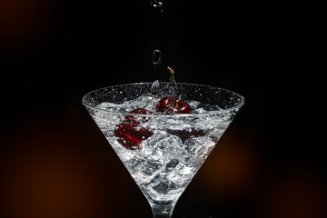 Close up view of splash water with falling cherry in a martini glass among ice in black background foreground.