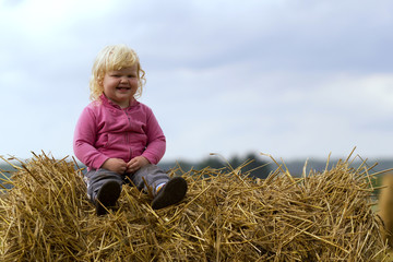 Naklejka na ściany i meble healthy childhood in harmony with nature - happy girl sits on a haystack in a wheat field