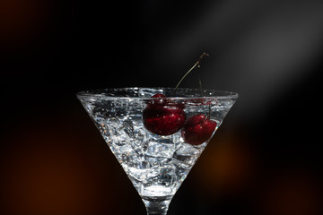 Close up view of splash water with falling cherry in a martini glass among ice in black background with flare and foreground.