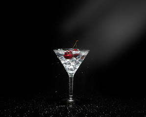 Close up view of splash water with falling cherry in a martini glass among ice in black background with flare.