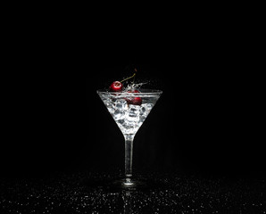 Close up view of splash water with falling cherry in a martini glass among ice in black background.