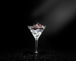 Close up view of splash water with falling cherry in a martini glass among ice in black background with flare.