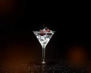 Close up view of splash water with falling cherry in a martini glass among ice in black background foreground.