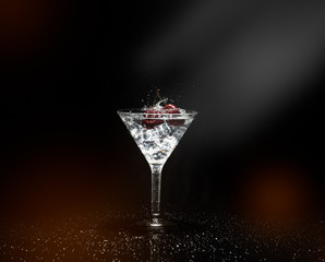 Close up view of splash water with falling cherry in a martini glass among ice in black background with flare and foreground.