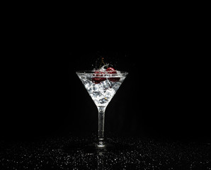 Close up view of splash water with falling cherry in a martini glass among ice in black background.