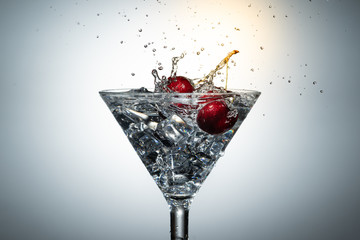 Close up view of splash water with falling cherry in a martini glass among ice in white background with flare .