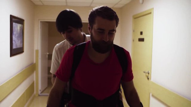 Portrait of young disable man walking in the robotic exoskeleton through the corridor of the rehabilitation clinic. Doctor walking behind him.