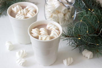 Cocoa with marshmelow in white cups and marshmallows in a glass jar on a white table with spruce branches and garland.