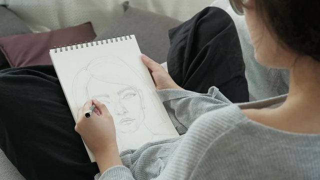 Young woman artist painting sketch on paper notebook with pencil. Woman sitting on sofa in living room, holding pencil and drawing sketch